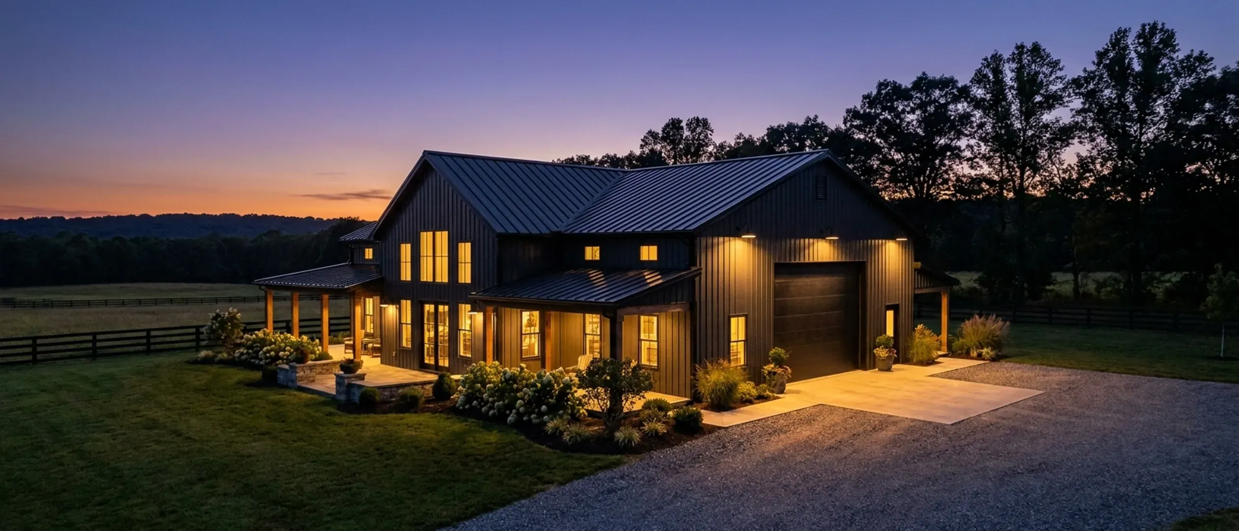 A barndominium at dusk with warm light glowing from every window and exterior lighting along the porch and roofline, rolling farmland and a low wooded ridge behind.