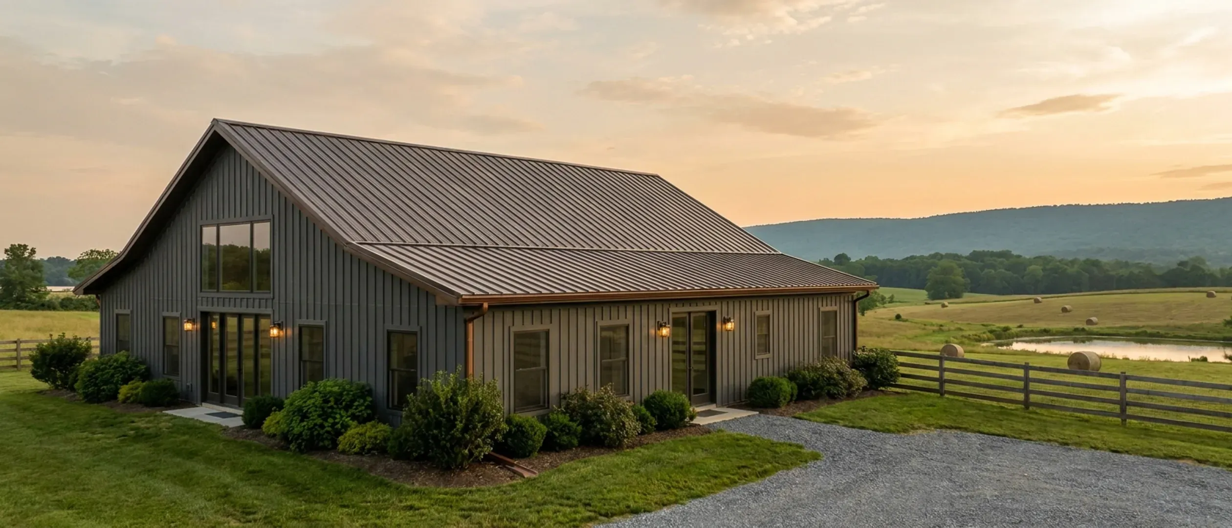 A completed barndominium viewed from a low angle showing its standing seam metal roof, vertical wall panels, gutters and exterior wall lights.